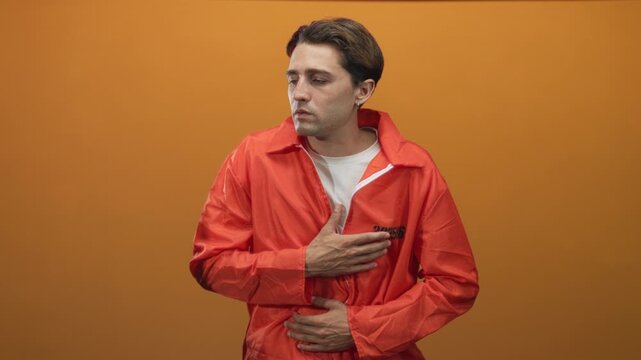 Man in orange jumpsuit with visible inmate number clutching chest with hand and holding stomach, white tee visible, studio backdrop; remorse vulnerability confinement.