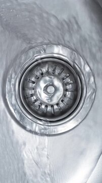 Water Flowing Into Sink Drain. Clear water swirls across a stainless steel basin and drains through a metal strainer.