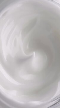 White Cream Swirling. Thick glossy cream moves in smooth folds, creating a soft abstract cosmetic texture.	