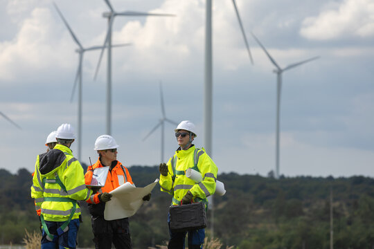 engineer and worker team stand in green field holding blueprint while discussing renewable wind energy project near tall turbine wearing protective safety hardhat on site