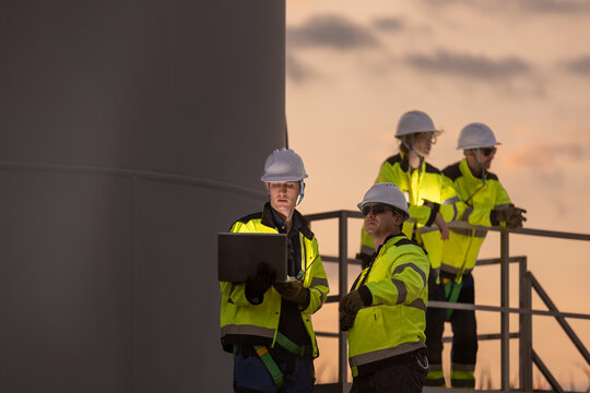 Group of confident industrial worker team wearing yellow reflective safety vest and white hard hat checking laptop computer outside large wind turbine during beautiful sunset