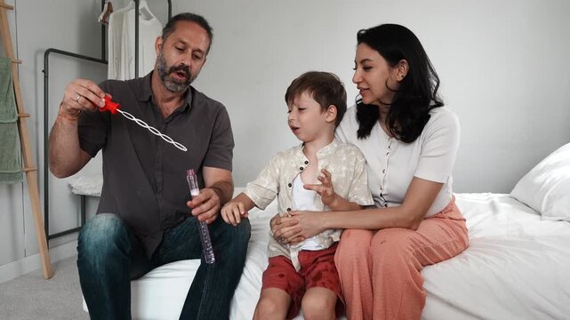 A boy, his mother, and his father sit together on the bed, playing with fun toys and holding a bubble blower at home. A family-themed concept.