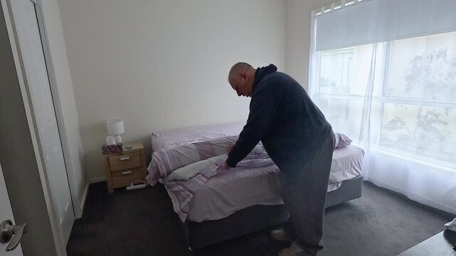 Middle aged man gets frustrated while trying to fit a doona cover over a duvet in a modest bedroom.