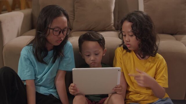 Three siblings, two little girls and a young boy, watch a tablet together on a living room sofa, capturing family bonding, children's screen time, home entertainment, and happy family moments.