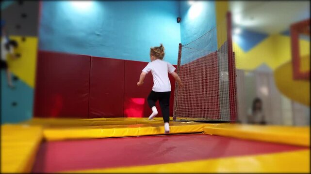 Young blonde child jumping with enthusiasm on indoor trampoline at entertainment center. Active little girl enjoying bouncy fun in a colorful play zone.