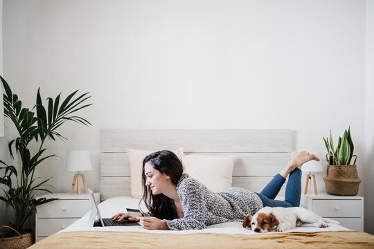 Woman with credit card doing online shopping through laptop while lying on bed by Jack Russell Terrior at home