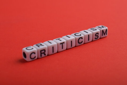 The word criticism spelled with alphabet cubes on a red background, symbolizing evaluation, judgment, feedback, opinion, communication, conflict, and constructive or negative review.