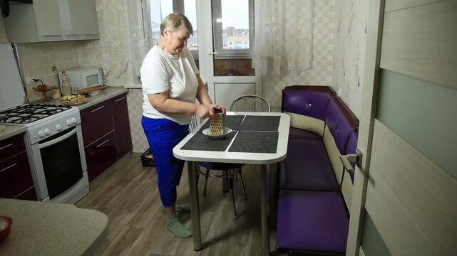 Elderly woman grating food at kitchen table, standing in home kitchen preparing ingredients for meal, domestic scene of everyday cooking activity