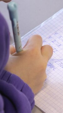 A student writing math equations in a grid notebook with a pen at a desk, capturing the essence of academic diligence and concentrated effort during a classroom study session. Vertical