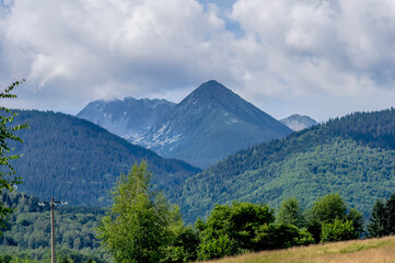 Obraz premium A panoramic mountain landscape featuring the majestic peak of Retezat Mountain in the Retezat National Park, Romania, framed by dense coniferous forests, lush green slopes, and a cloudy blue sky.
