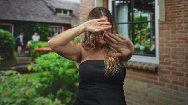 Woman shields eyes with hands and holds palms forward in front of brick building window and garden; shyness privacy solitude.