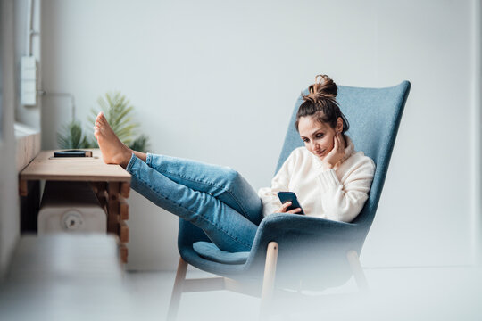 Young woman using smart phone sitting in armchair at home