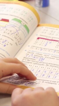 A student solves mathematics homework in an open workbook at a desk, capturing the process of learning arithmetic and basic geometry concepts in a bright classroom setting. Vertical