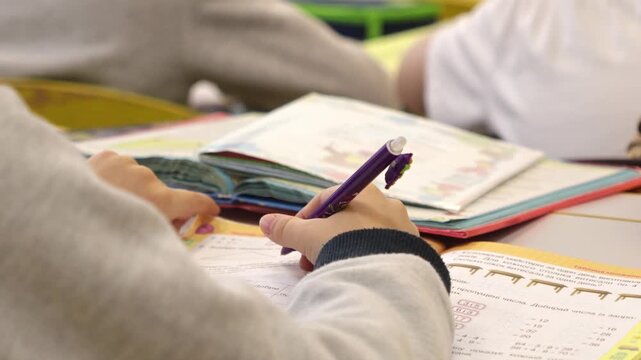 A student using a pen to solve math problems in a workbook at a desk, representing the focus, concentration, and academic progress of a young learner during a classroom study session