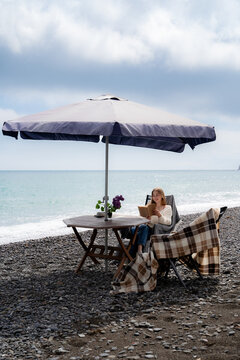Minimal seaside composition with a woman relaxing under a beach table with umbrella on a rocky coastline. Mindfulness, leisure and coastal lifestyle.