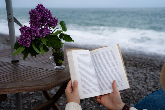 Close-up of hands holding an open paper book by the sea. Relaxing coastal reading, mindfulness and slow living.