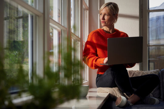 Mature woman sits indoors by window with laptop feeling peaceful