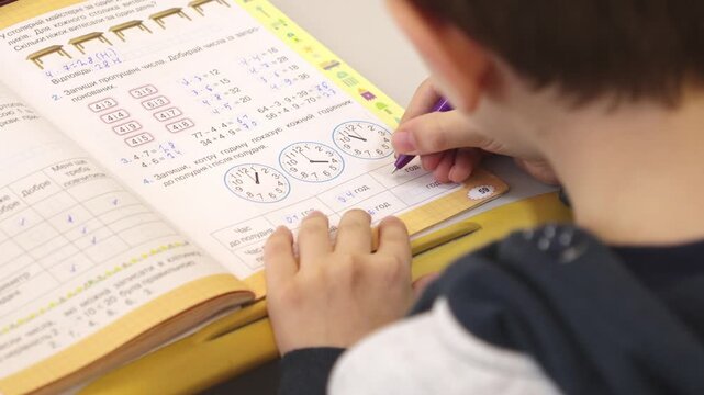 A young student diligently solves math problems in a workbook while sitting at a desk, demonstrating deep concentration and dedication during an elementary academic lesson today