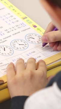 A young student writing answers in a math workbook at school desk, capturing the effort and concentration involved in learning time telling skills during a daily lesson. Vertical