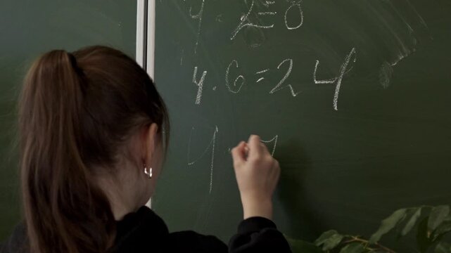 A student writing mathematical equations on a green classroom chalkboard with chalk, representing the process of academic learning and engagement in a school education environment now