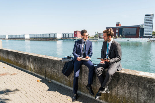 Two young businessmen sitting on wall by river, working