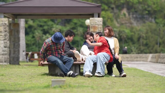 Five friends relax on a park bench. They chat and laugh, sharing joyful moments. Green grass and trees frame their cozy gathering. Ideal for scenes of friendship, leisure, or outdoor bonding