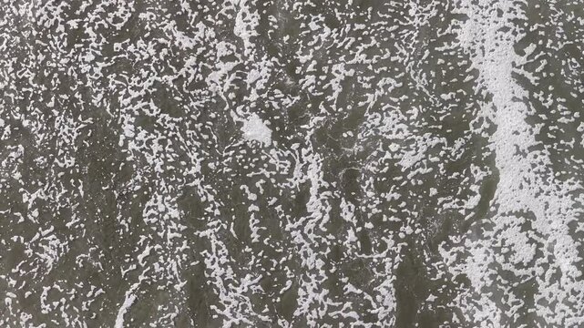 Drone topdown view of white foam waves receding on black volcanic sand from directly above, moody nature and dramatic coastal view.