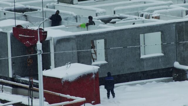 Construction workers clearing snow from building foundation on cold winter day; crane equipment on site; snowy weather conditions; labor work outdoors; development project