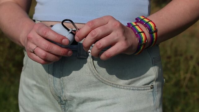 Hands of a girl in light jeans with rainbow bracelets and headphones in a case. On the girl's wrist are two fabric bracelets with a rainbow print LGBTQ+. Hands with rainbow pride bracelets close-up