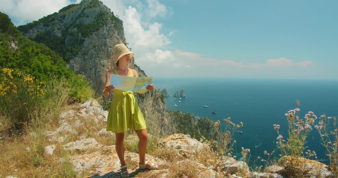 Woman holding a map on a rocky Capri cliff overlooking the sea and distant Faraglioni stacks