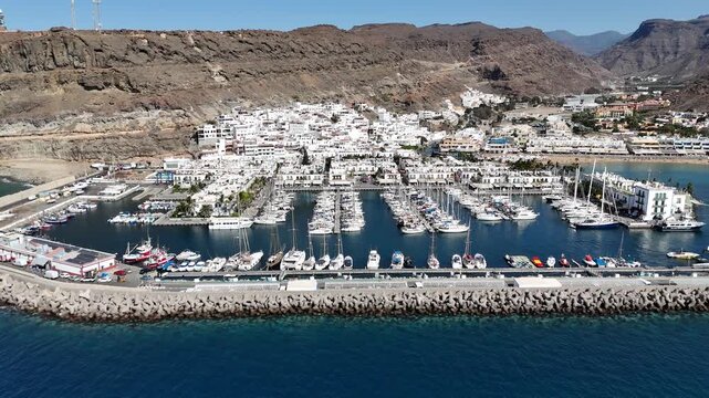 Pan shot of marina with sailboats at Atlantic ocean in Puerto de Mogan, white houses and beach with mountains in the background on Canary Island Gran Canaria, Spain