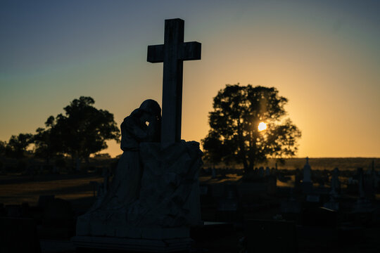 Silhouette of a grieving angel statue at a cemetery during sunrise