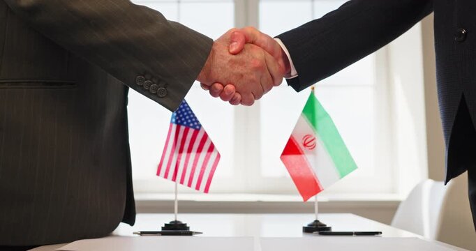 Iran USA flags handshake diplomacy agreement negotiation. Two suited diplomats shake hands at a table as flags signal politics and treaty talks. Strong symbol of international diplomatic accord.