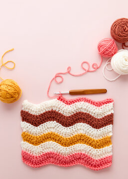 Colorful crochet project with yarn balls and crochet hook on pastel pink background. DIY crochet project with colorful wool and hook, flat lay.