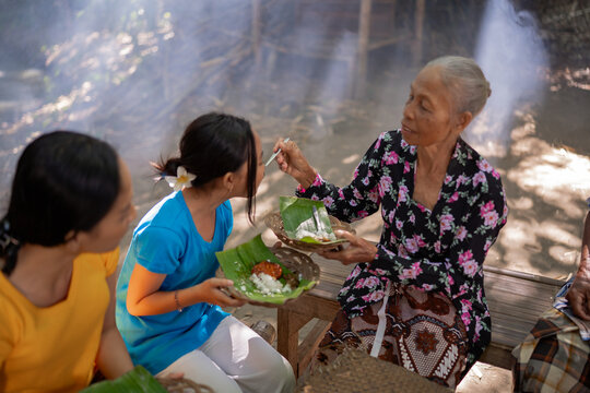 Older woman feeds younger women during outdoor meal, holding banana leaf plates with rice and side dishes on rustic benches, with soft sunlight, haze, and relaxed family atmosphere in natural setting