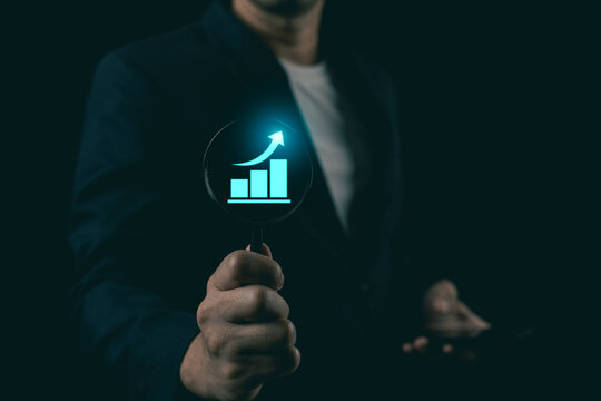 Close up of businessman holding magnifying glass over glowing business growth graph, concept of financial analysis and market research.