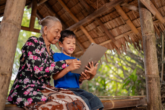 Smiling elderly woman guides young boy using tablet inside rustic bamboo shelter, captured in warm natural daylight with close family interaction, intergenerational support, and simple rural living