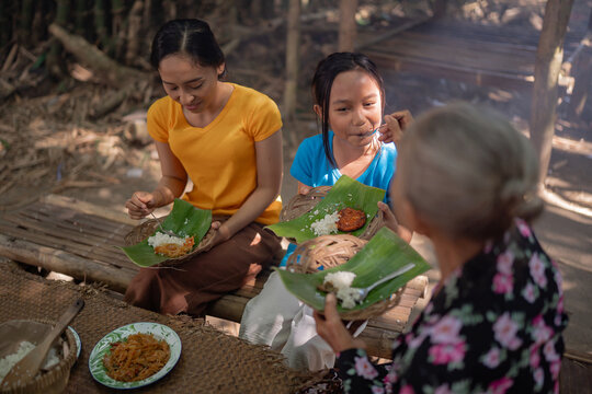 Two women and young girl eating homemade food from banana leaf plates on woven baskets beside rustic bamboo seating, capturing family style dining, traditional food culture, and warm outdoor rural