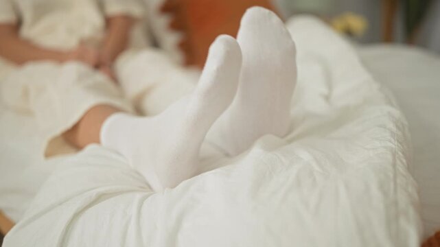 Middle aged blonde woman lying on a bed with white socks, feet raised on a rumpled duvet in a cozy bedroom set; relaxation comfort.