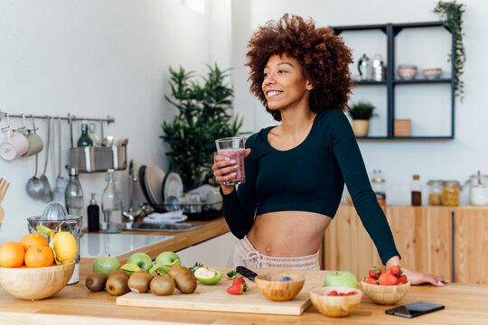 Smiling woman with Afro hairstyle holding glass of smoothie standing in kitchen