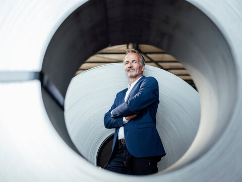 Confident businessman with arms crossed standing by metal sheet roll