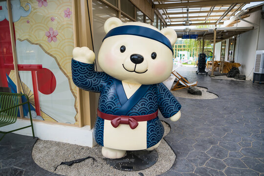 Bear Mascot in Traditional Japanese Attire: A cheerful bear mascot, dressed in a blue happi coat and headband with a wave pattern, stands welcomingly outdoors.