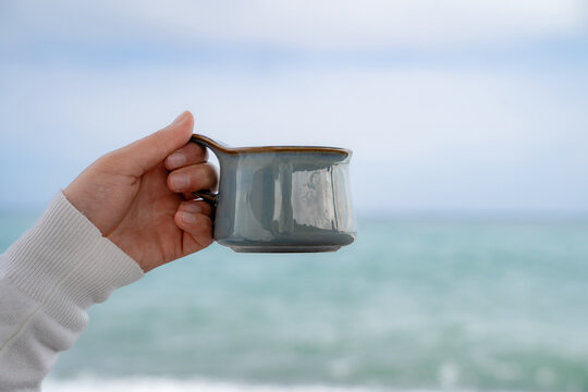 Close-up of a woman&rsquo;s hand holding a ceramic cup with the ocean in the background. Mindful relaxation, coastal lifestyle and cozy outdoor moment.