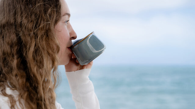 Young curly woman enjoying a warm drink while looking at the ocean. Slow living, peaceful seaside atmosphere and outdoor wellbeing concept.