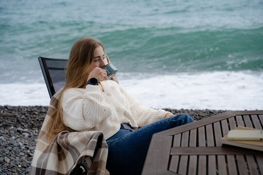 Young woman warming up with a hot drink by the ocean. Mindful relaxation, seaside leisure and calm travel concept.