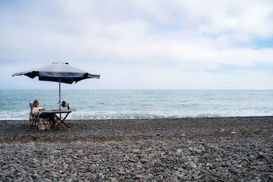 A woman spends time alone, looking at the sea horizon, sitting at a table while enjoying the view of nature. Mindfulness, outdoor recreation, coastal mental recovery, and solitude.