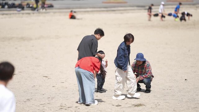 Sunlit beach with families and children playing. People lean over sand, laughing and interacting. One child crouches nearby, curious and engaged. Ideal for family outings or beach holiday themes