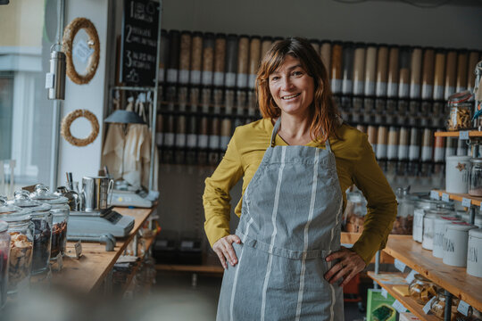 Smiling female entrepreneur in apron with hand on hip standing in zero waste shop