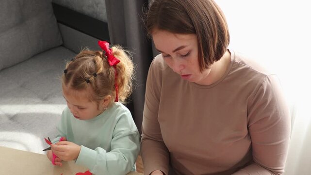 Portrait of a young Caucasian mother and her little daughter making paper crafts and hearts together at home from a top view. Family creativity, child education, parenting, and emotional bonding conce