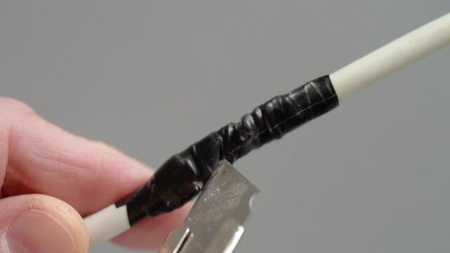 Person using utility knife to strip tape from electrical wire. Close perspective of hands carefully removing tape from white electrical cable with utility knife